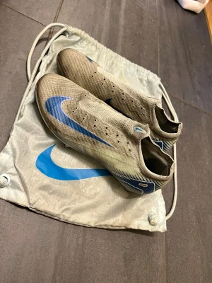Nike mercurual vapor 14 elite   blå - Säljer ett par Nike fotbollsskor i  blå detaljer och stor Swoosh-logga på sidan. Skorna har en strömlinjeformad design, rund tå och är tillverkade i syntetmaterial för lätt känsla på planen. Perfekta för dig som vill sticka ut på matchen. Skolåda följs givetvis med