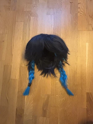 Svart-blå peruk med flätor - Venti peruk. Från Genshin Impact, med svart hår och två långa blå flätor. Håret är rakt och har en lite rufsig look upptill, medan flätorna är tydligt markerade och hålls ihop med svarta och blå hårband. Perfekt för cosplay eller maskerad…. 