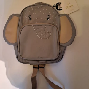 Grå elefant-ryggsäck från Rofa Design - Supergullig grå ryggsäck från Rofa Design, formad som en elefant med stora öron och broderat ansikte. Väskan har beige detaljer, justerbara axelband och dragkedja framtill. Perfekt för dig som vill ha en unik och lekfull accessoar.