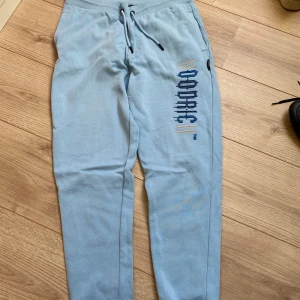 Ljusblå mjukisbyxor med tryck - Snygga ljusblå mjukisbyxor med snörning i midjan och ett coolt blått tryck på ena benet. Byxorna har en avslappnad passform och är perfekta för chill dagar. Mjukt material och elastisk midja för extra komfort.