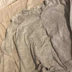 Grå kofta från H&M med knappar - Mysig grå kofta från H&M i storlek 134/140. Koftan har rund halsringning, långa ärmar och stängs med knappar framtill. Tillverkad i mjuk bomullsmix och passar perfekt till lager-på-lager under höst och vinter.