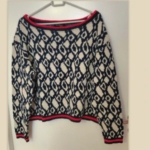Mönstrad stickad tröja Fuchsia London, M - Stickad tröja med ett mönster i nyanser av mörkblått och beige. Tröjan har en bred halsringning med en röd detalj och matchande detaljer vid ärmslut och nederkant. Fint skick.  Storlek M.  Från armhålan till armhålan 58cm, längd 51cm.