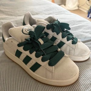 Adidas Campus vita och gröna sneakers - Säljer ett par Adidas Campus sneakers i vitt mocka med gröna detaljer och breda gröna snören. Det finns också vita snören till ni kan få med om jag hittar dem😅 Knappt använda, kanske typ 3 gånger. Har sedan bara stått i garderoben. Lite noppriga i hälarna bara och lite smuts på baksidan men knappast märkbart. 