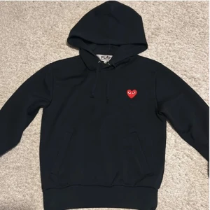 Svart hoodie Comme des Garçons Play - Svart hoodie från Comme des Garçons Play med huva och snörning. Klassisk röd hjärtlogga med ögon på bröstet. Mjuk bomullskänsla, känguruficka framtill och ribbade muddar. Bara och skriva om minsta fundering!