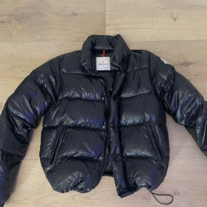 Svart Moncler pufferjacka - Svart pufferjacka från Moncler med glansig finish och klassisk logga på ärmen. Jackan har hög krage, två dragkedjefickor och snörning nedtill. Fylld med äkta dun för extra värme. Perfekt för dig som vill ha en snygg och ikonisk vinterjacka öppen för byten! .