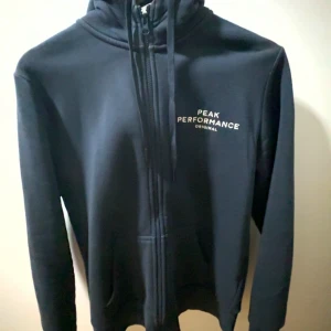 Svart hoodie från Peak Performance - Svart hoodie från Peak Performance med dragkedja, huva och snörning. Logga på bröstet och ärmen. Mjuk insida och klassisk passform, perfekt för chill eller träning.