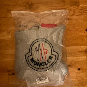 Grå Moncler hoodie med dragkedja - Snygg grå hoodie från Moncler med hel dragkedja, huva med snörning och stora loggan framtill. Röd och vit detalj på huvan och ärmslut. Mjuk bomullskvalitet och klassisk passform, perfekt för en chill och sportig look.