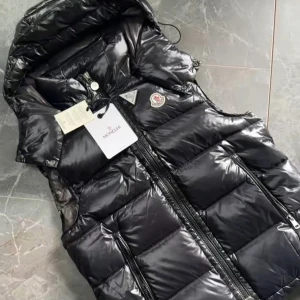 Svart dunväst från Moncler med huva - Snygg svart dunväst från Moncler med glansig finish och quiltad design. Västen har en stor avtagbar huva, två dragkedjeförsedda fickor och Moncler-logga på bröstet. Perfekt för lager-på-lager och streetstyle. Dragkedja framtill och cool puffer-look.