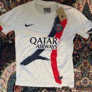 PSG tröja - Snygg vit Paris Saint-Germain fotbollströja från Nike med klubbmärke på bröstet och Qatar Airways-tryck. Tröjan har korta ärmar och coola penseldrag i blått och rött framtill. Perfekt för dig som älskar fotboll och vill sticka ut på planen.