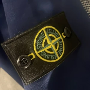 Blå vindjacka från Stone Island - Säljer en blå vindjacka från Stone Island med huva och hel dragkedja framtill. Jackan har den klassiska Stone Island-loggan på vänster ärm och är tillverkad i ett slätt, lätt material. Perfekt för dig som vill ha en stilren och sportig look.