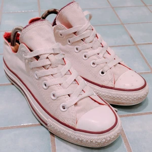 Vita Converse Chuck Taylor All Star Sneakers [39] - Klassiska vita Converse Chuck Taylor All Star sneakers i canvas med low top och röda detaljer. Skorna har snörning, rund tå och platt sula. Perfekta för dig som gillar en tidlös och avslappnad stil. Knappt använda men finns en liten fläck på hättan på en av skorna. Se bilder. 