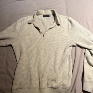 Beige stickad half zip-tröja Riley - Säljer en beige stickad tröja från Riley med krage och half zip-dragkedja. Tröjan har lång ärm och en snygg, diskret struktur i materialet. Perfekt för lager-på-lager och chill stil.