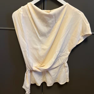 Champagnefärgad blus från SHEIN - Snygg champagnefärgad blus från SHEIN i storlek S. Blusen har korta ärmar, omlott-detalj med knyt i sidan och ett lätt glansigt material som ger en lyxig känsla. Perfekt för dig som gillar en feminin och trendig look.