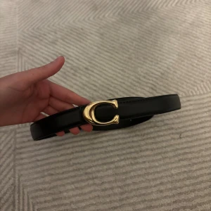 Svart skärp - Coach med gulddetalj - Snyggt svart skärp Coach inspirerad med ett stilrent guldfärgat C-spänne framtill. Bältet är gjort i slätt skinn och passar perfekt till jeans eller klänning. 