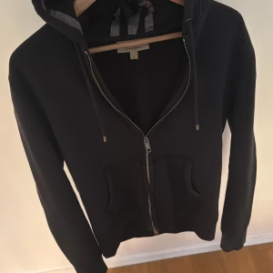 Svart hoodie från Burberry med dragkedja - Mörkblå hoodie från Burberry med klassiskt rutigt foder i huvan, hel dragkedja framtill och två fickor. Snygg och enkel design med långa ärmar och justerbar snörning i huvan. 