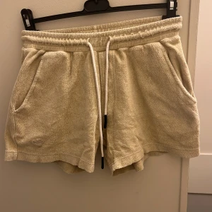 Beige frottéshorts från OAS - Säljer ett par beige shorts från OAS i mjuk frotté-bomull. De har elastisk midja med snörning, två sidofickor och en bakficka. Perfekta för sommardagar och chill vibes. Klassisk och avslappnad passform.