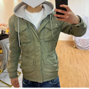Grön fieald jacket från j lindeberg  - Snygg militärgrön parkajacka från J Lindeberg  med fyra stora fickor framtill, hög krage och dragkedja. Jackan har en klassisk patch på ärmen och är tillverkad i polyester. Perfekt för dig som vill ha en stilren och funktionell jacka o glida med stil!  Obs ej jag på första bilden modellen är 175!