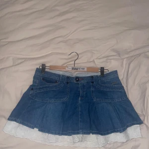 Jeanskjol med spets från BDG Urban Outfitters - Supersöt blå jeanskjol från BDG Urban Outfitters med volangig vit spetskant nedtill. Kjolen har klassiska jeansfickor, bälteshällor och knappar framtill. Perfekt för dig som gillar denim med en extra touch av romantisk stil.