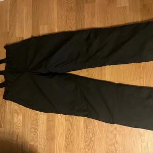 Svarta täckbyxor Neomondo Junior 164cl - Svarta täckbyxor från Neomondo med justerbara hängslen och diskret logga på benet. Perfekta för vinterns alla äventyr, tillverkade i slitstarkt syntetmaterial som står emot kyla och snö. Snygg och enkel design som funkar till allt.