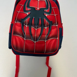 3D  Spiderman ryggsäck - Cool ryggsäck med Spiderman-tema i rött och blått. Framsidan har ett stort svart spindelmotiv och mönster som liknar ett spindelnät. Väskan har röda axelband och blå baksida, perfekt för dig som gillar superhjältar.