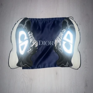 Svarta Dior B30 sneakers med reflex - Säljer ett par svarta Dior B30 sneakers med vita detaljer och reflekterande CD-logga på sidorna. Skorna har tjock, vit och svart platt sula, rund tå och vita snören. Materialet är en mix av syntet och mesh som ger en sportig vibe. Perfekt för dig som gillar exklusiva och trendiga sneakers.