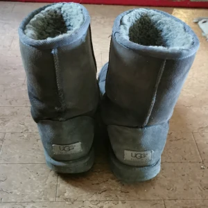 Grå UGG boots i mocka - Mysiga grå boots från UGG i mjuk mocka med fluffigt foder på insidan. Klassisk rund tå och platt sula, perfekta för kalla dagar. UGG-logga baktill på hälen. Enkla att dra på och superbekväma.