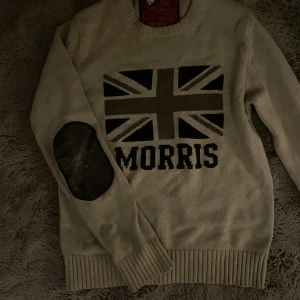 Beige stickad tröja Morris - Beige stickad tröja från Morris med Union Jack-flagga och texten 'Morris' framtill. Tröjan har svarta armbågslappar och ribbade muddar vid ärmslut och nederkant. Perfekt för dig som gillar preppy stil och snygga detaljer.