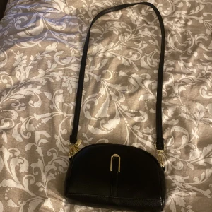 Svart väska - En svart väska med guldiga detaljer. Aldrig använd. Vet tyvärr inte märke på den. Man kan byta band på den . Den har två fack och ett litet till fack. 