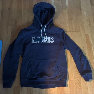 Marinblå hoodie från Morris, storlek L - Säljer en marinblå hoodie från Morris i storlek L. Tröjan har vit snörning i huvan, stor ficka framtill och tydlig Morris-logga på bröstet. Perfekt för dig som gillar en klassisk och stilren stil. 