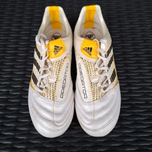Adidas Predator fotbollsskor vit/gul - Adidas Predator fotbollsskor i vitt och gult med svarta detaljer. Skorna har snörning, platt sula med dobbar och klassisk rund tå. Snyggt rutigt mönster på sidan och Predator-logga på tungan. Tillverkade i skinn för grym komfort och känsla på planen.