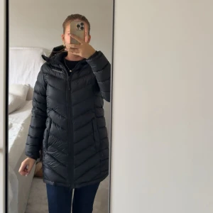 Svart lång dunjacka Peak Performance  - Svart lång dunjacka från Peak Performance med huva och dragkedja framtill. Jackan har tyvärr en defekt på ena armen men inget som syns när man har på sig den! Perfekt till hösten/vinter. Köpt för 5000kr