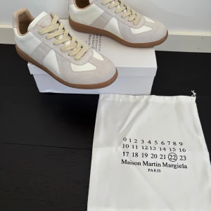 Maison Margiela sneakers - Maison Margiela sneakers i beige och vitt med klassisk gummisula. Ovandel i skinn och mocka, beige snörning och rund tå. Snygg minimalistisk design med diskreta detaljer och ikonisk Margiela-logga på plösen. Perfekta för dig som gillar stilrena och trendiga sneakers.