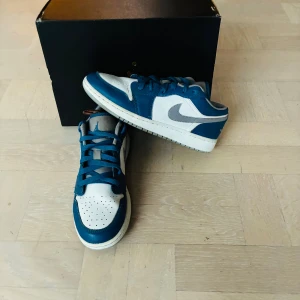 Nike Jordan 1 LOW TRUE BLUE CEMENT(GS) - Nike Jordan 1 Low sneakers i blått och vitt med grå swoosh. Skorna har snörning, perforerad tå och platt sula. Klassisk rund tå och lågt skaft. Materialet är skinn och syntet, vilket ger en snygg och sportig look. Perfekta för dig som gillar streetwear och sneakers. Priset är inte hugget i sten!