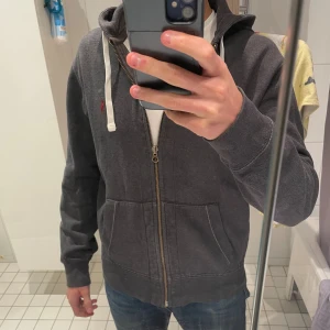 Mörkgrå zip up hoodie från Polo Ralph Lauren - Mörkgrå hoodie från Polo Ralph Lauren med dragkedja, vita snören och klassisk röd logga på bröstet. Tröjan har huva, kängurufickor och är tillverkad i mjuk bomullsmix. Perfekt för chill dagar eller när du vill ha en clean streetstyle. True to size, flitigt använd och tidlös kvalitet! Axelbredd 47 och längd 62, passar svin bra på 175-185 cm långa personer