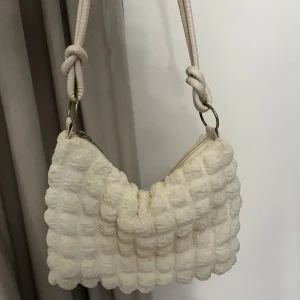 Vit quiltad axelväska med struktur - . Väskan har en justerbar axelrem med knutdetaljer och stängs med dragkedja upptill. Perfekt för att lyfta vilken outfit som helst. Man får plats med super mycket saker