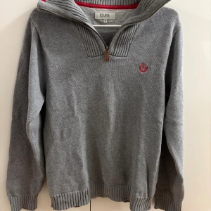 Grå stickad tröja från Etirel - Snygg grå stickad tröja från Etirel med ribbade muddar och krage. Tröjan har en half zip-dragkedja och ett rött broderat märke på bröstet. Perfekt för kyliga dagar och enkel att matcha med jeans eller chinos.