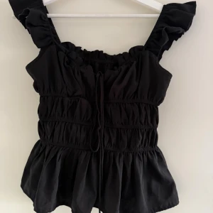 Svart volangig blus med knytband - Supersöt svart blus med volangiga axelband och smockade detaljer. Toppen har knytband framtill och en luftig, rynkad passform som ger en romantisk vibe. Perfekt för dig som gillar feminina detaljer och vill sticka ut lite extra.