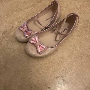 Rosa ballerinaskor med rosett från Graceland - Söta ballerinaskor i ljusrosa från Graceland med glansig finish och perforerat mönster. Skorna har rund tå, dekorativ rosett framtill och justerbar rem runt vristen. Perfekta för dig som gillar en feminin och lekfull stil.