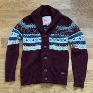 Mörkröd kofta från Superdry med mönster - Säljer en sjukt snygg mörkröd kofta från Superdry med nordiskt mönster i vitt, blått och grått. Koftan har stora knappar, bred krage och är stickad i mjukt material. Perfekt för kalla dagar och ger en chill vibe till din outfit. Modellen är 176 och den passar bra på han.