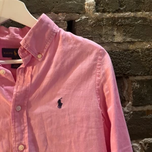 Rosa skjorta från Ralph Lauren - Klassisk rosa skjorta från Ralph Lauren med button-down krage och broderad marinblå logga på bröstet. Skjortan har långa ärmar, knappar framtill och är tillverkad i mjuk bomull och linne. Perfekt för en stilren och avslappnad look.