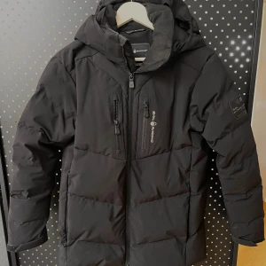 Svart fown jkt från Sail Racing - Svart jacka från Sail Racing med huva och flera dragkedjeförsedda fickor. Jackan är vadderad, har en stilren look och är tillverkad i syntetmaterial med DWR-behandling. Perfekt för kalla dagar och har logga på bröstet och ärmen.