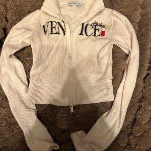 Vit ribbad kofta från Bershka - Vit ribbad kofta från Bershka med broderad text 'Venice Italy' och en liten italiensk flagga framtill. Tröjan har lång ärm, hög krage och dragkedja hela vägen. Perfekt för lager-på-lager och snygg till jeans eller kjol.