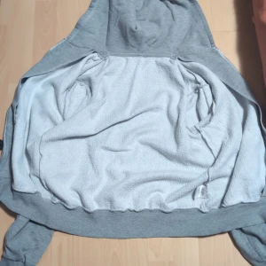 Grå hoodie med dragkedja och huva - Säljer en enkel grå hoodie med dragkedja och huva. Tröjan har långa ärmar, fickor framtill och ett broderat nummer på bröstet. Perfekt för chill dagar eller när du vill ha något bekvämt och stilrent. Materialet är mjukt och känns skönt mot huden.