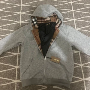 Grå zip up hoodiejacka från Burberry Med dragkedja  - Säljer en grå hoodiejacka från Burberry med klassiskt rutigt foder i huvan. Jackan har dragkedja framtill, ribbade muddar och två fickor. Perfekt för dig som gillar stilrena och exklusiva plagg med en twist av ikoniskt mönster.