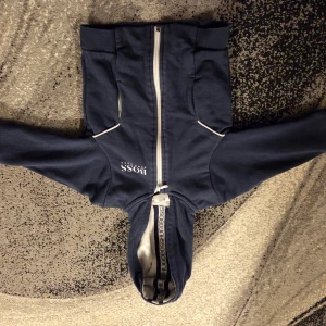 Marinblå hoodie från Hugo Boss - Snygg marinblå hoodie från Hugo Boss med vit dragkedja och logga på bröstet. Tröjan har huva med logotypband på insidan och två fickor framtill. Perfekt för dig som vill ha en stilren och sportig look.