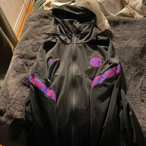 Svart Nike FC Barcelona vindjacka - Svart zip från Nike med FC Barcelona-logga på bröstet och blå-röda detaljer längs ärmarna. Jackan har huva med snörning och hel dragkedja framtill. Perfekt för dig som vill visa ditt stöd för Barca och samtidigt hålla stilen.