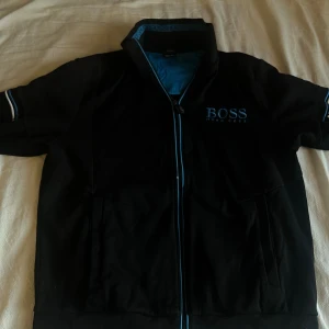 Svart Boss zip up  - Svart track jacket från Hugo Boss med blå detaljer och broderad logga på bröstet. Jackan har dragkedja framtill, ståkrage och ribbade muddar. Tillverkad i mjuk bomullsmix, perfekt för en sportig och clean look.