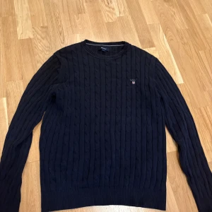 Mörkblå kabelstickad tröja från GANT - Säljer en mörkblå kabelstickad tröja från GANT med broderad logga på bröstet. Tröjan har rund halsringning, ribbade muddar och är tillverkad i mjuk bomull. Perfekt för dig som gillar klassisk och stilren look. Men har tyvärr en defekt vid kragen vilket sänker mitt pris