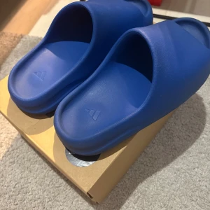 Adidas Yeezy Slide Azure blå - Säljer ett par Adidas Yeezy Slide i färgen Azure. Slidesen är helblå, har en chunky och modern design med räfflad sula och öppen tå. Tillverkade i syntetmaterial för maximal komfort och stilpoäng. Perfekta för chill dagar eller när du vill sticka ut.