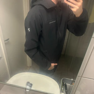 Svart vindjacka Peak Performance - Säljer en svart vindjacka från Peak Performance med huva och dragkedja framtill. Jackan har diskreta loggor på bröstet och ärmen samt justerbar huva. Perfekt för blåsiga dagar och enkel att matcha med jeans eller träningskläder.